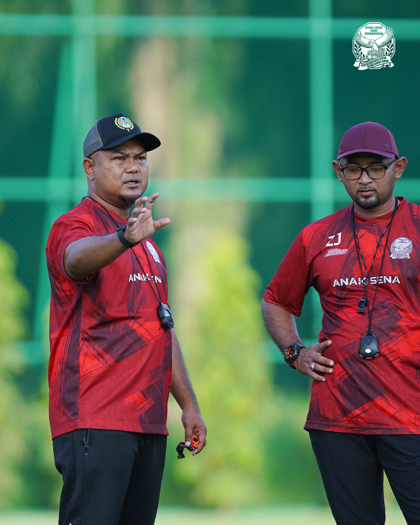 Akmal Rizal in a red Kedah training shirt and cap directing a session, with a member of his coaching staff alongside him