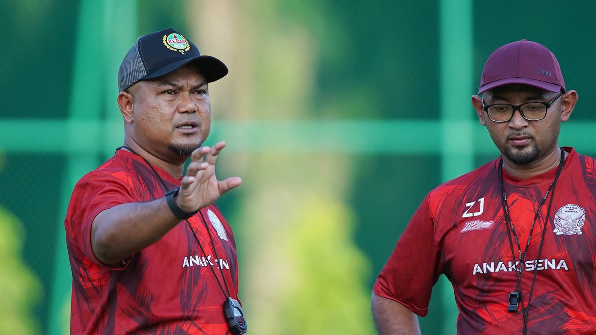 Akmal Rizal in a red Kedah training shirt and cap directing a session, with a member of his coaching staff alongside him